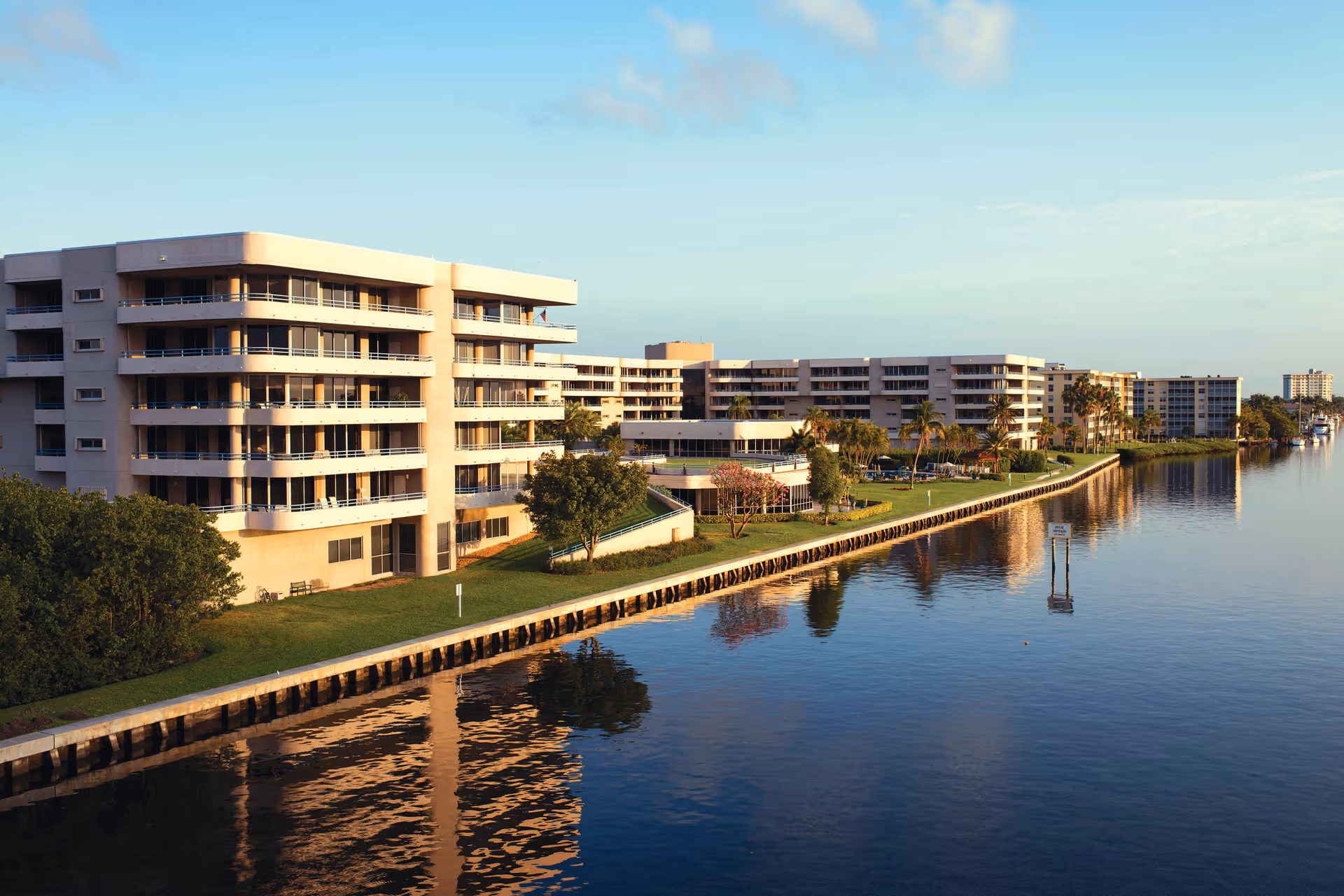 Multi-story waterfront residential buildings with balconies and palm trees reflected in a calm waterway.