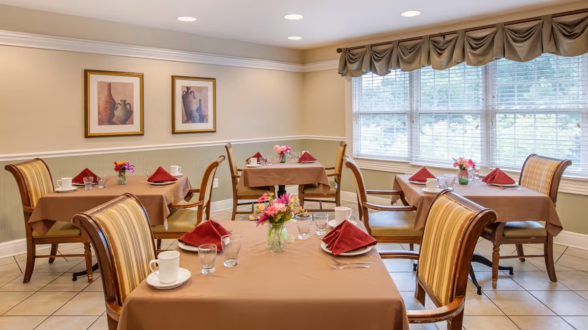 A bright dining room with four tables covered in brown tablecloths, each set with red folded napkins, white cups, glasses, and small flower arrangements. The room has large windows with blinds and valances, beige walls with white trim, and two framed pictures on the wall.