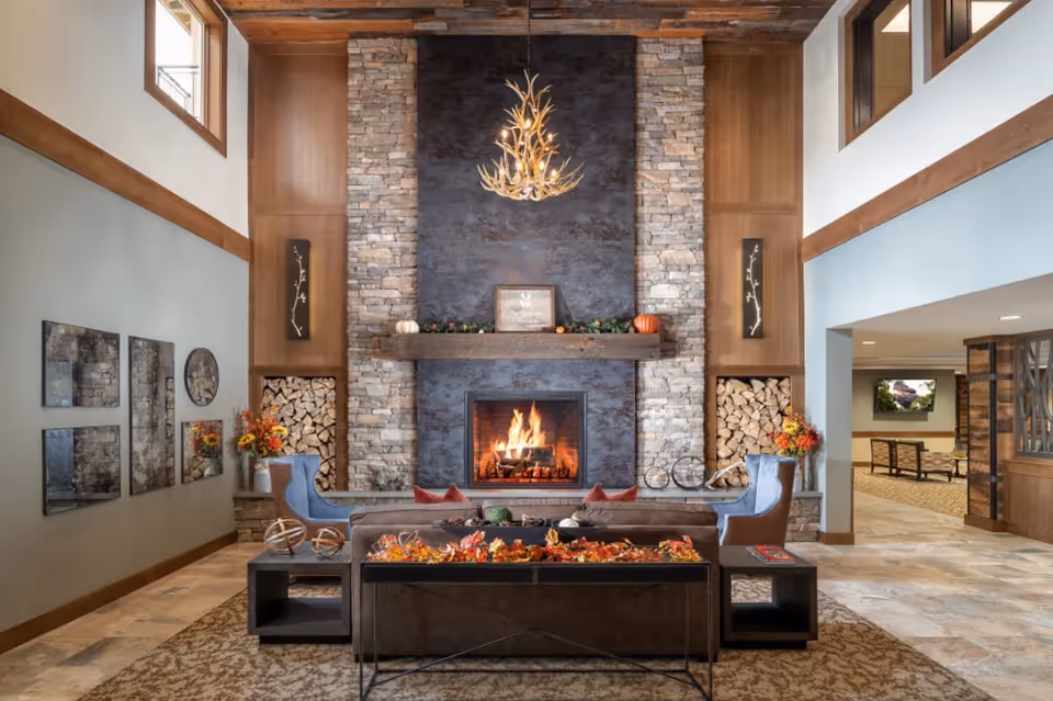 A cozy living room area with a large stone fireplace in the center, decorated with autumn-themed items on the mantel and a fire burning inside. Two blue armchairs flank the fireplace, and a brown sofa faces it with a table behind it adorned with fall leaves and decorative spheres. The room features high ceilings with wooden beams, windows near the ceiling, and a chandelier made of antlers hanging above the fireplace. The walls have wood paneling and stone accents, and there are decorative mirrors and flower arrangements on the left wall.