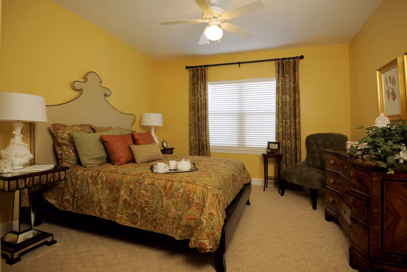 A warmly decorated bedroom with yellow walls, a large bed with an ornate headboard, patterned bedspread and multiple pillows. There are two bedside tables with lamps, a window with closed blinds and patterned curtains, a green upholstered chair, and a wooden dresser with plants and framed artwork above it.