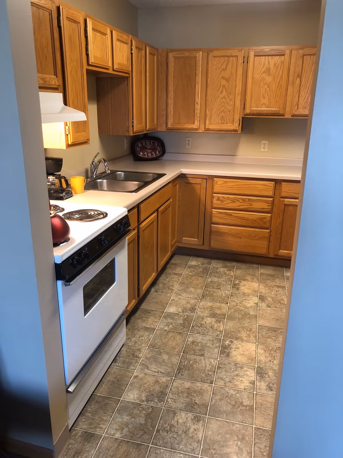 A small kitchen with wooden cabinets, a white electric stove with a red kettle on it, a double stainless steel sink, a coffee maker, and a clock on the countertop. The floor is covered with brown and gray patterned tiles.