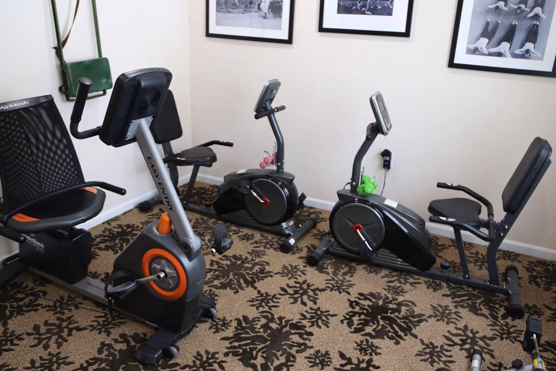 A small exercise room with three recumbent stationary bikes placed on a patterned carpet. The walls are light-colored and decorated with three black and white framed photos of people exercising. There is a green bag hanging on the wall in the corner.