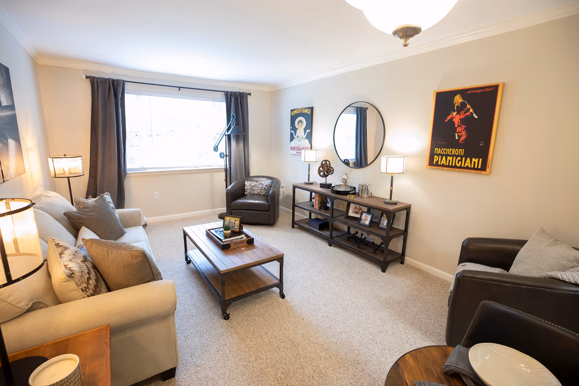 A bright and cozy living room with a beige sofa adorned with multiple pillows, two dark leather armchairs, a wooden coffee table with books and decor, and a console table against the wall with lamps, framed photos, and decorative items. The room has a large window with dark curtains, light-colored walls, and two vintage-style posters hanging on the wall.