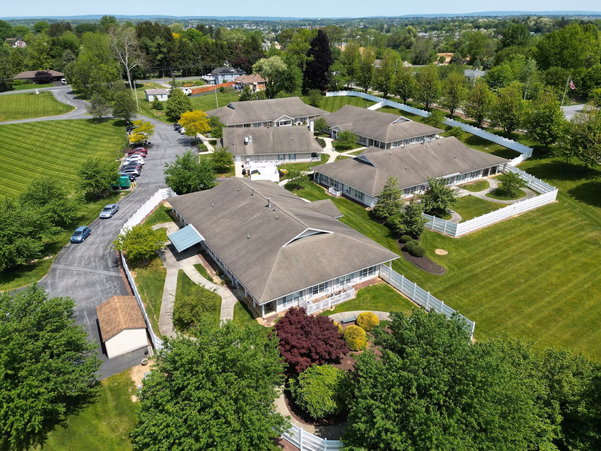 Aerial view of Linden Village A ProMedica Senior Living Community showing multiple single-story buildings with gray roofs surrounded by green lawns, trees, and white fences. There is a paved driveway with parked cars and a small shed near the entrance. The surrounding area includes more greenery and distant residential houses under a clear sky.