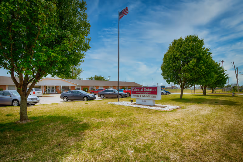 Exterior view of Shawnee Colonial Estates nursing and rehabilitation facility with a lawn, trees, parked cars, and an American flag on a flagpole in front of the building under a partly cloudy sky.