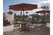 Outdoor patio area with several metal tables and chairs, each shaded by large red umbrellas. Palm trees and other greenery are visible in the background under a partly cloudy sky.