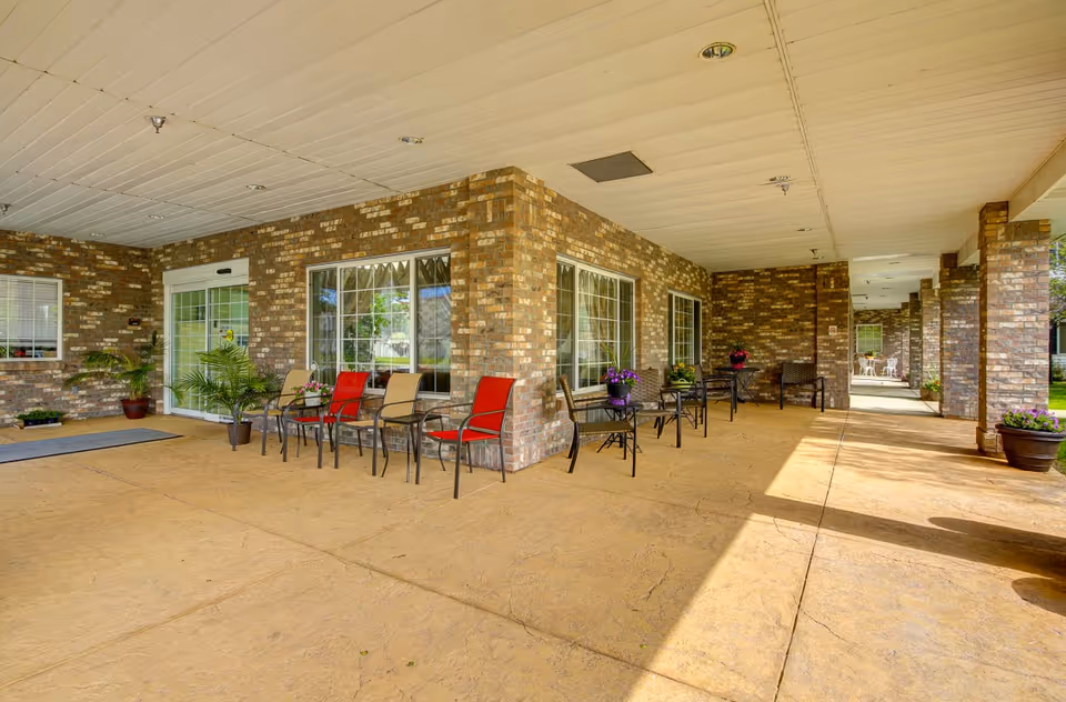 Covered brick entrance patio with chairs and small tables arranged along a walkway at an assisted living facility.