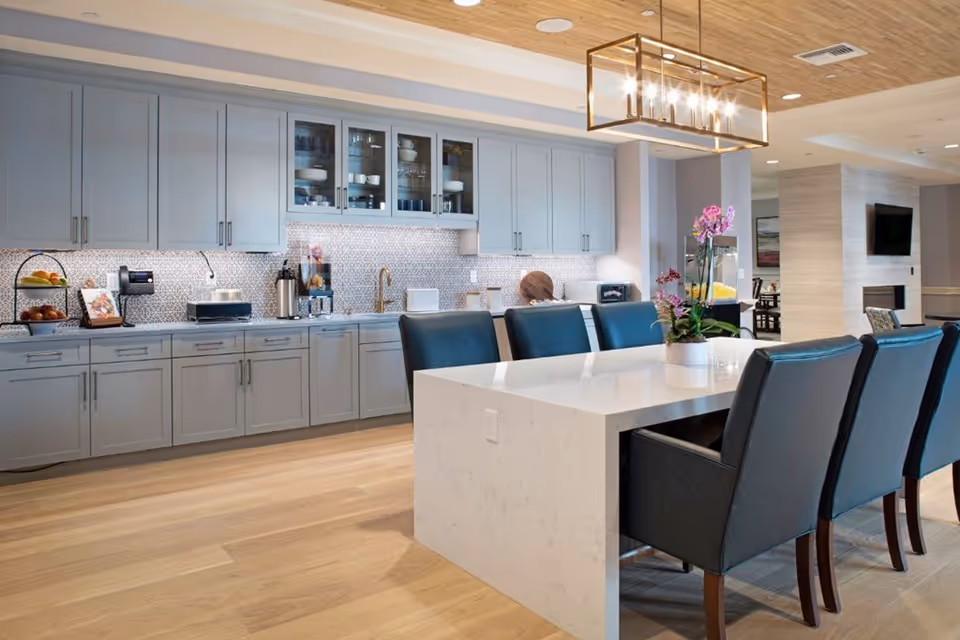 Modern kitchen area with light gray cabinets, a white marble island with black cushioned chairs, a decorative light fixture above the island, and a backsplash with a geometric pattern. The space has wooden flooring and a ceiling with wooden paneling. There is a small plant with purple flowers on the island and a popcorn machine in the background.