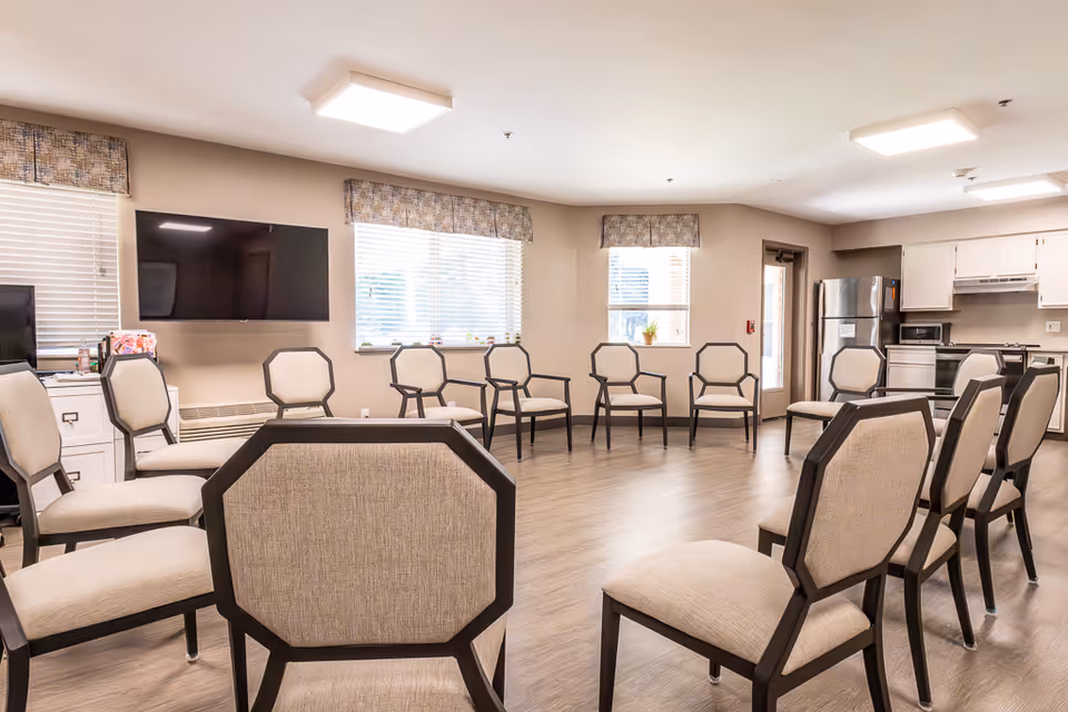 A spacious, well-lit common room with beige walls and wood flooring, featuring a circle of cushioned chairs with dark wooden frames. The room includes large windows with patterned valances, a wall-mounted flat-screen TV, and a kitchen area with white cabinets, a stainless steel refrigerator, and a stove in the background.