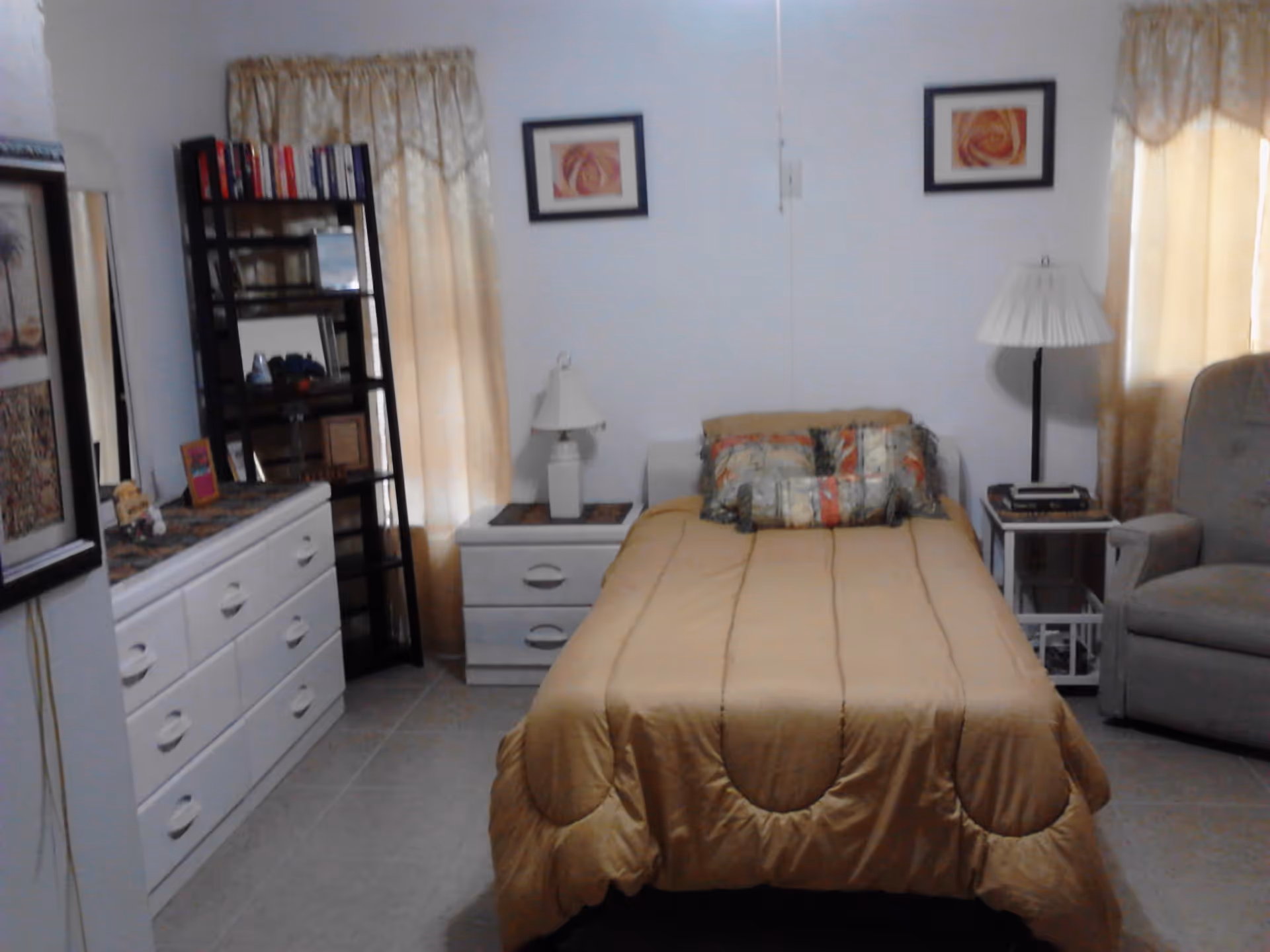 A bedroom with a single bed covered with a gold-colored comforter and decorative pillows. There are two framed pictures on the wall above the bed. On either side of the bed are white nightstands with lamps. To the left is a white dresser with multiple drawers and a black shelving unit with books and decorative items. Beige curtains cover the windows, and a gray armchair is positioned to the right of the bed.