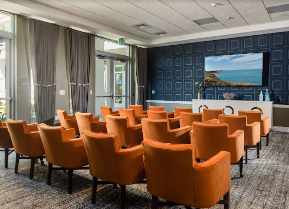 A room with multiple orange upholstered chairs arranged in rows facing a wall-mounted flat screen TV displaying a coastal scene. The room has large windows with gray curtains, a patterned dark blue accent wall behind the TV, and a light-colored cabinet with decorative items underneath the TV.