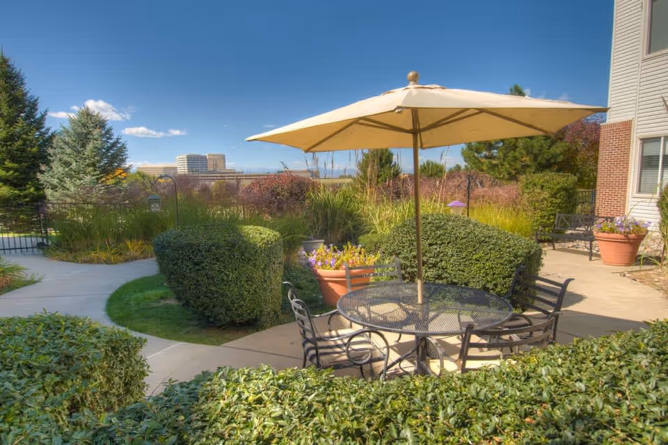 Outdoor patio area at Caley Ridge Assisted Living featuring a round metal table with a large beige umbrella and four chairs. The patio is surrounded by well-maintained bushes, potted plants, and trees, with a clear blue sky and buildings visible in the background.