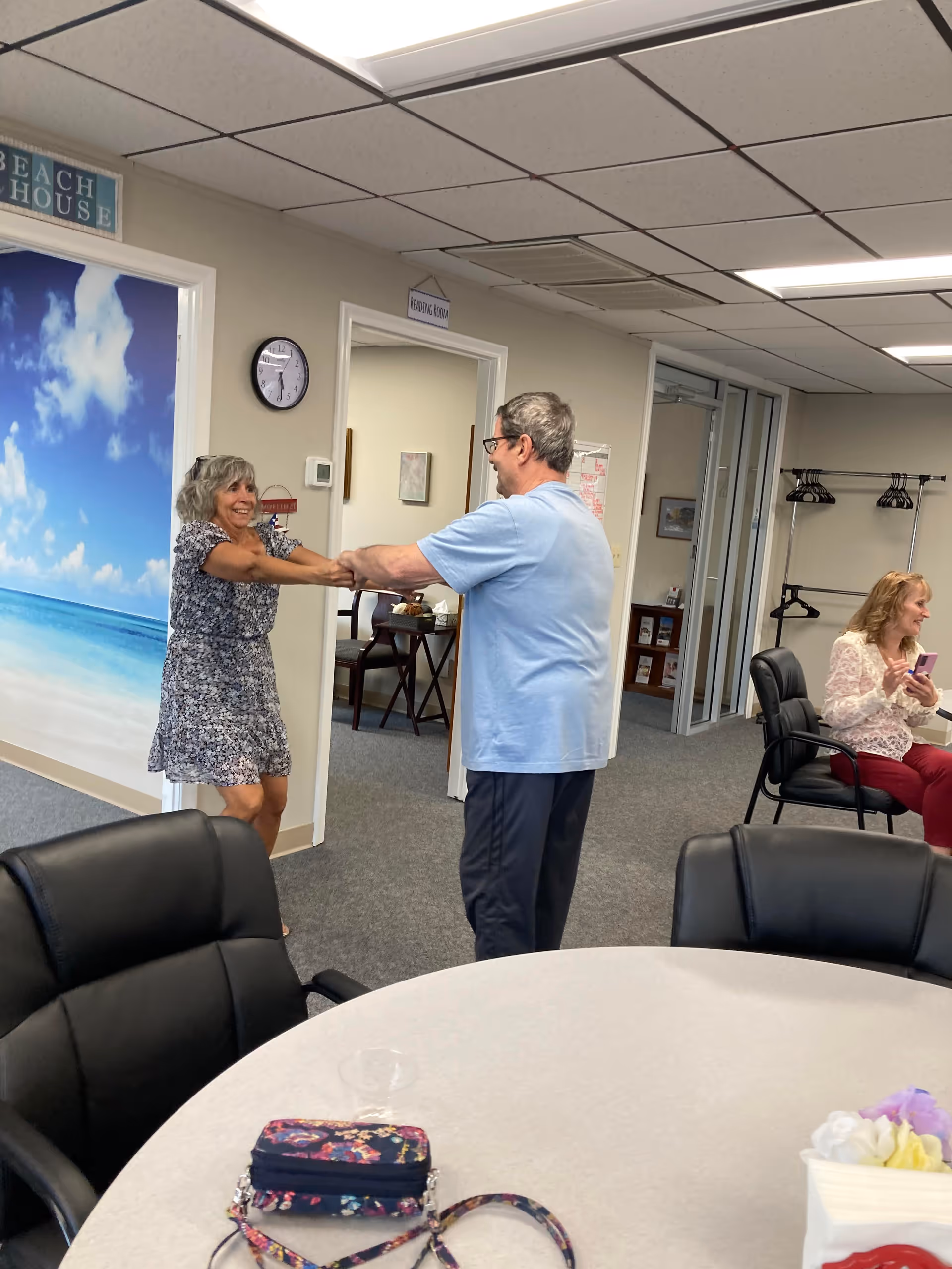 Two older adults holding hands and smiling while standing in an indoor room with a beach-themed wall mural. Another woman is sitting on a chair nearby, looking at her phone. The room has office chairs, a round table with a floral purse on it, and a coat rack in the background.