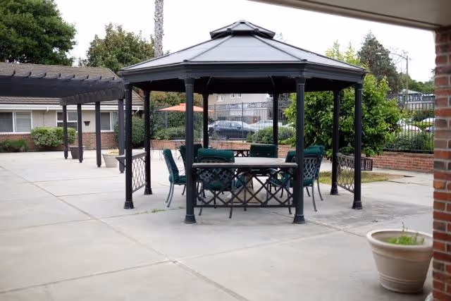 Outdoor patio area with a black metal gazebo covering a round table surrounded by green cushioned chairs. The patio is paved with concrete and has a pergola structure on the left side. There are bushes, trees, and a fence in the background along with a building and parked cars.