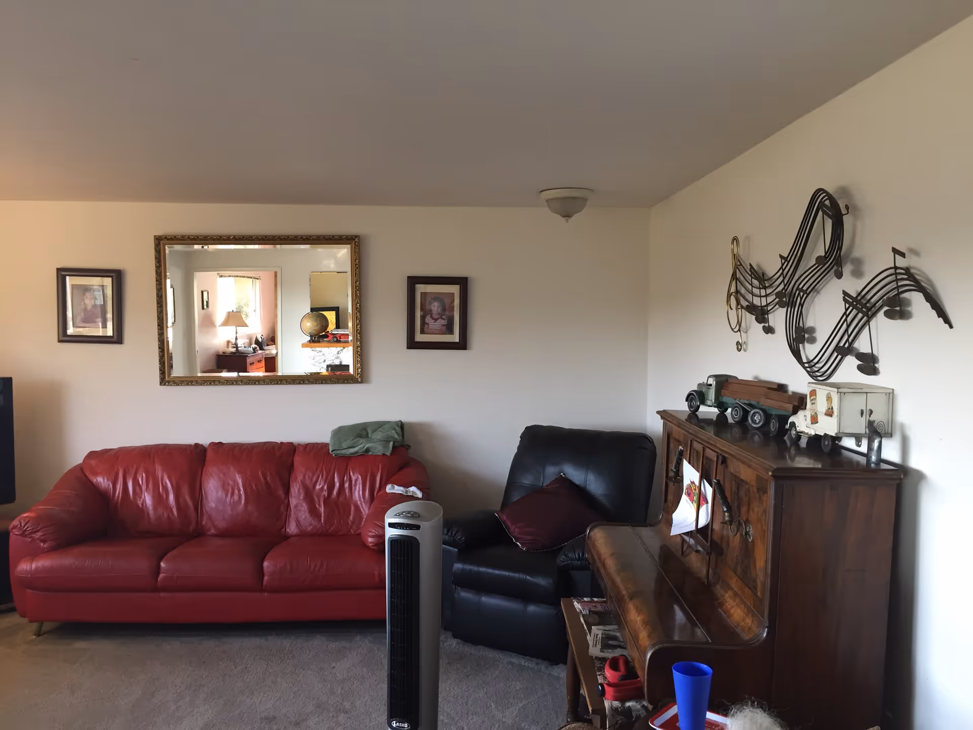 A living room with a red leather sofa and a black leather recliner chair. Above the sofa are two framed pictures and a large mirror reflecting another room. To the right, there is a wooden piano with decorative toy trucks on top and a musical note wall decoration above it. A tall, slim fan stands in front of the sofa on a carpeted floor.