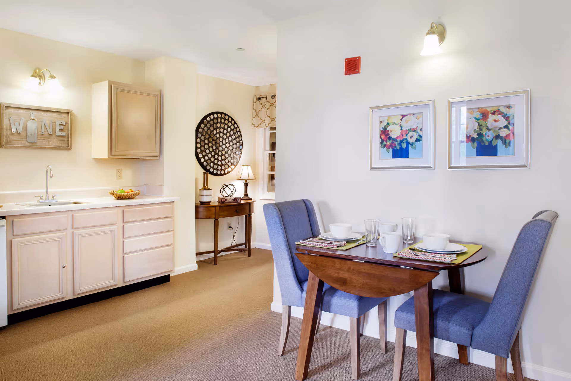 A small dining area with a wooden table set for two with white bowls, mugs, and glasses. Two blue upholstered chairs are placed at the table. Adjacent to the dining area is a kitchenette with light-colored cabinets, a sink, and a decorative sign on the wall that reads 'WINE'. The walls are painted white, and there are two framed floral paintings above the dining table. A small wooden side table with a lamp and decorative items is visible in the background.