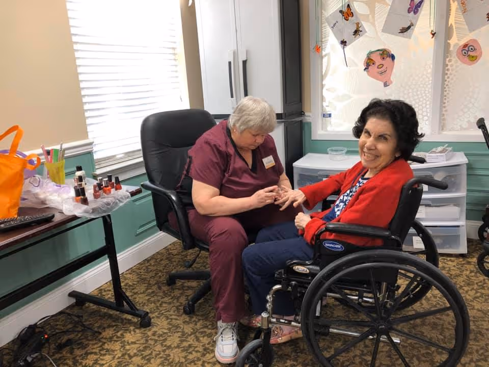 A caregiver in maroon scrubs is painting the nails of a smiling elderly woman seated in a wheelchair inside a room decorated with colorful paper crafts on the window. There is a table nearby with nail polish bottles and other manicure supplies.