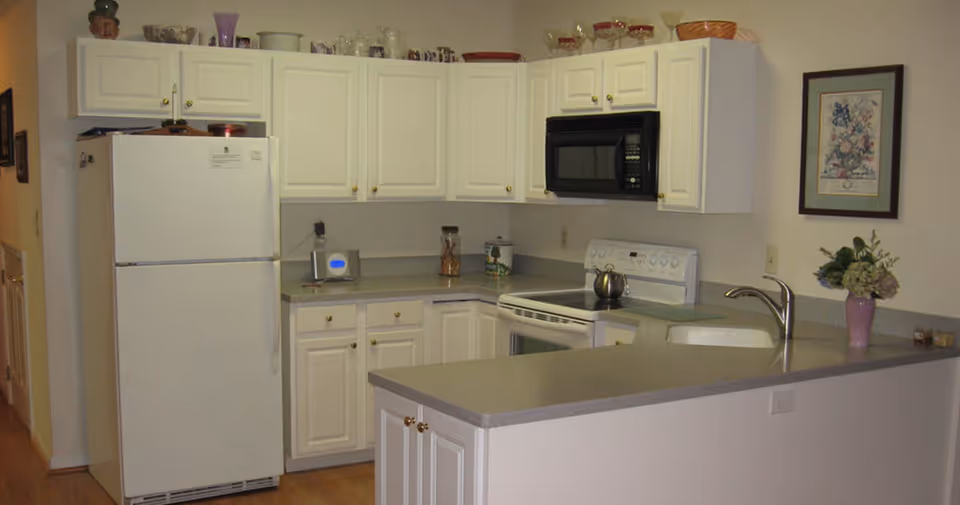 A kitchen with white cabinets and a light gray countertop. The kitchen includes a white refrigerator, a white electric stove with a black microwave above it, and a stainless steel sink with a faucet. There are various decorative items on top of the cabinets and a framed floral picture on the wall. A small vase with flowers is placed on the counter.