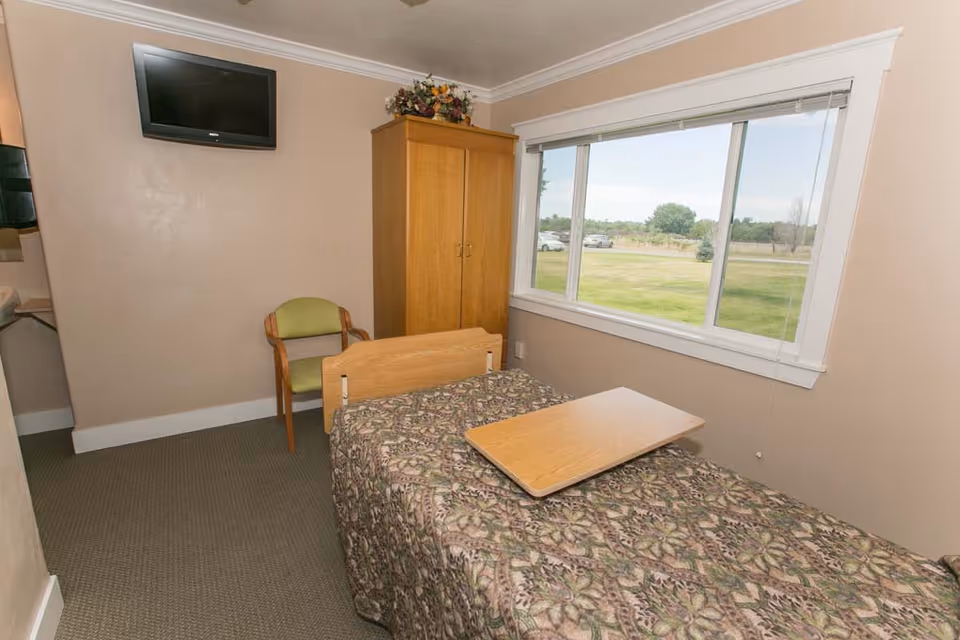 A small room with a single bed covered with a patterned bedspread and a wooden overbed table. There is a green cushioned wooden chair next to a wooden wardrobe with a floral arrangement on top. A flat-screen TV is mounted on the wall above the chair. A large window shows a grassy outdoor area with trees and parked cars.