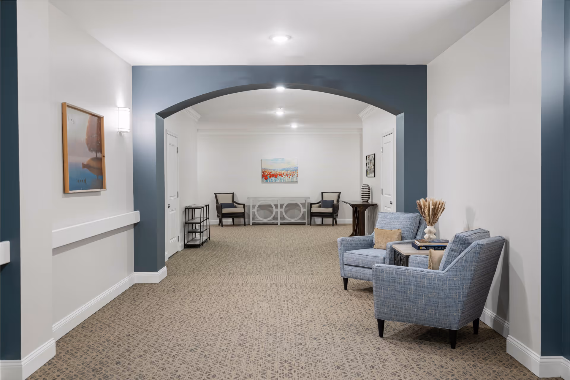 A spacious, well-lit interior hallway or common area with beige patterned carpet and white walls accented by a blue archway. Two blue upholstered armchairs with beige cushions are placed on the right side, accompanied by a small table holding books and a decorative vase with dried plants. At the far end, there are two dark wooden chairs with cushions, a white console table, and a colorful painting on the wall. Additional decorative elements include a framed picture on the left wall and a small black metal shelf unit.