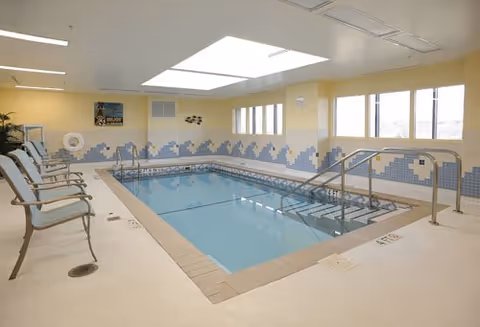 Indoor swimming pool with light blue water surrounded by beige tiled floor and walls decorated with blue and white tile patterns. Several chairs are lined up along one side of the pool. Large windows let in natural light and a skylight is visible on the ceiling.
