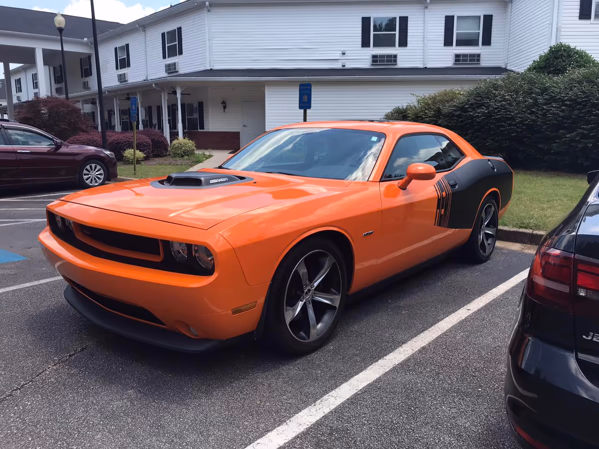 Orange and black Dodge Challenger parked in a parking lot in front of a white senior living building with black shutters and bushes nearby.