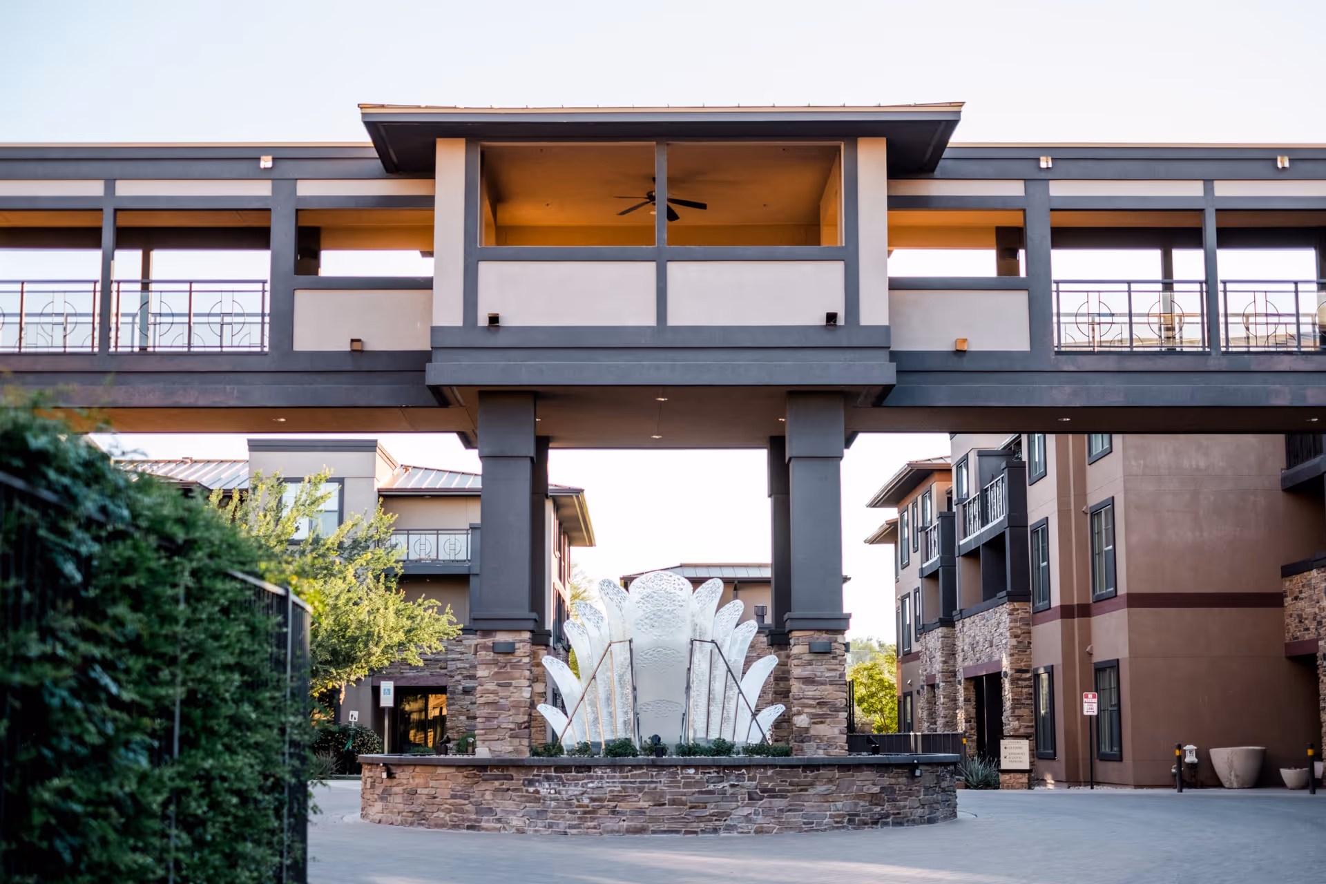 Exterior view of Andara By Cogir senior living facility featuring a modern architectural entrance with stone pillars and a decorative water fountain in the center, surrounded by multi-story buildings and greenery.