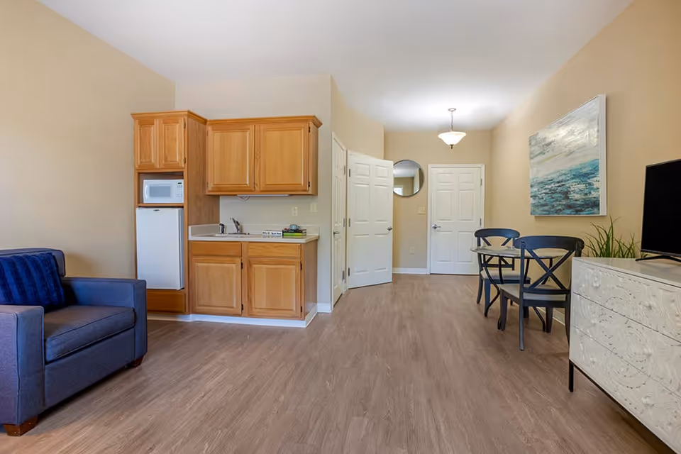 Interior view of a senior living facility room featuring a small kitchenette with wooden cabinets, a microwave, and a mini refrigerator. To the left is a blue armchair with a matching pillow. On the right side, there is a small dining table with two chairs, a decorative plant, a flat-screen TV on a white dresser, and a large abstract painting on the wall. The room has light-colored walls, wood flooring, and two white doors in the background.