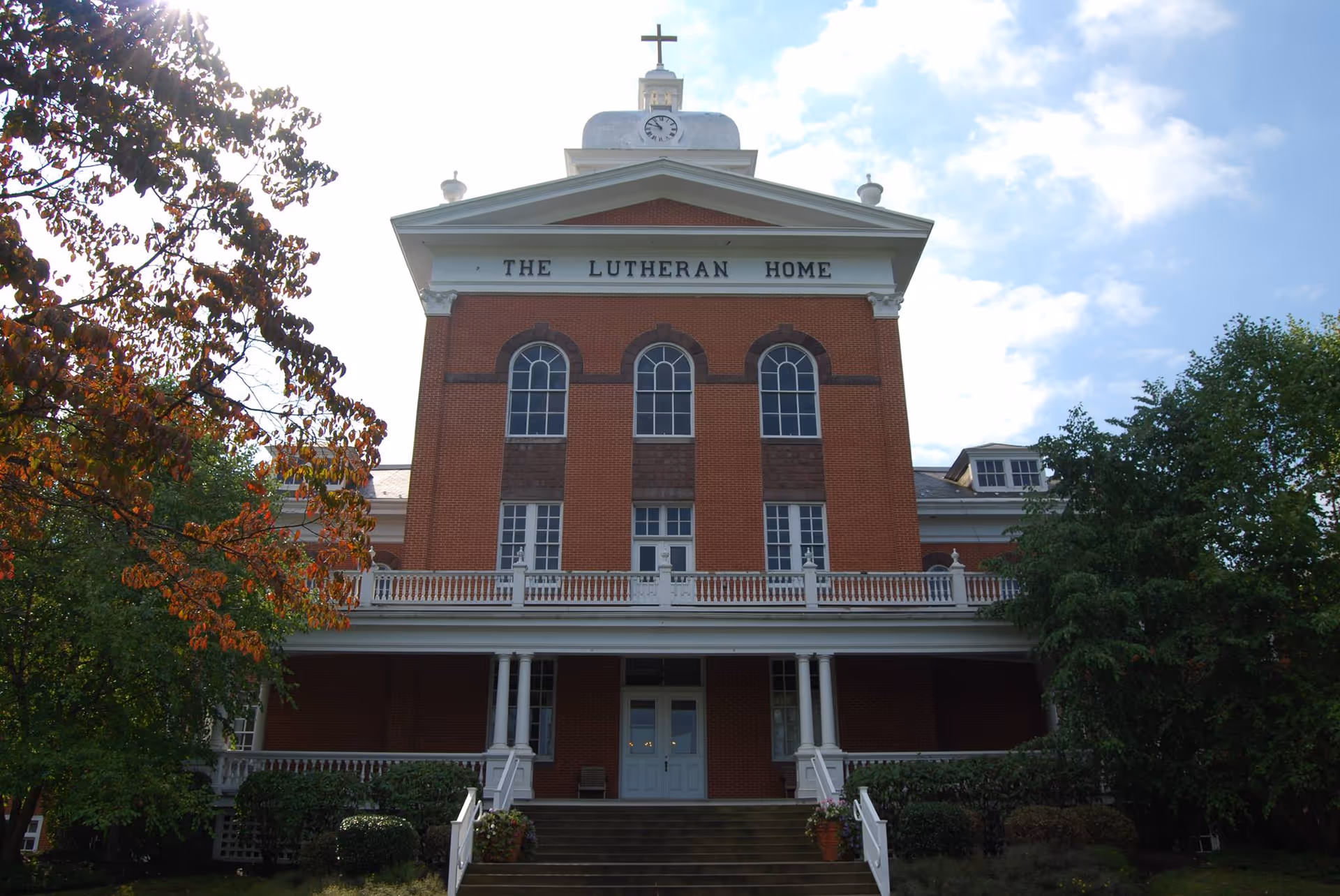Front exterior view of a large brick building with white trim and a cross on top. The building has three tall arched windows and a sign that reads 'THE LUTHERAN HOME'. There are trees with green and autumn-colored leaves on either side of the building and steps leading up to the entrance.
