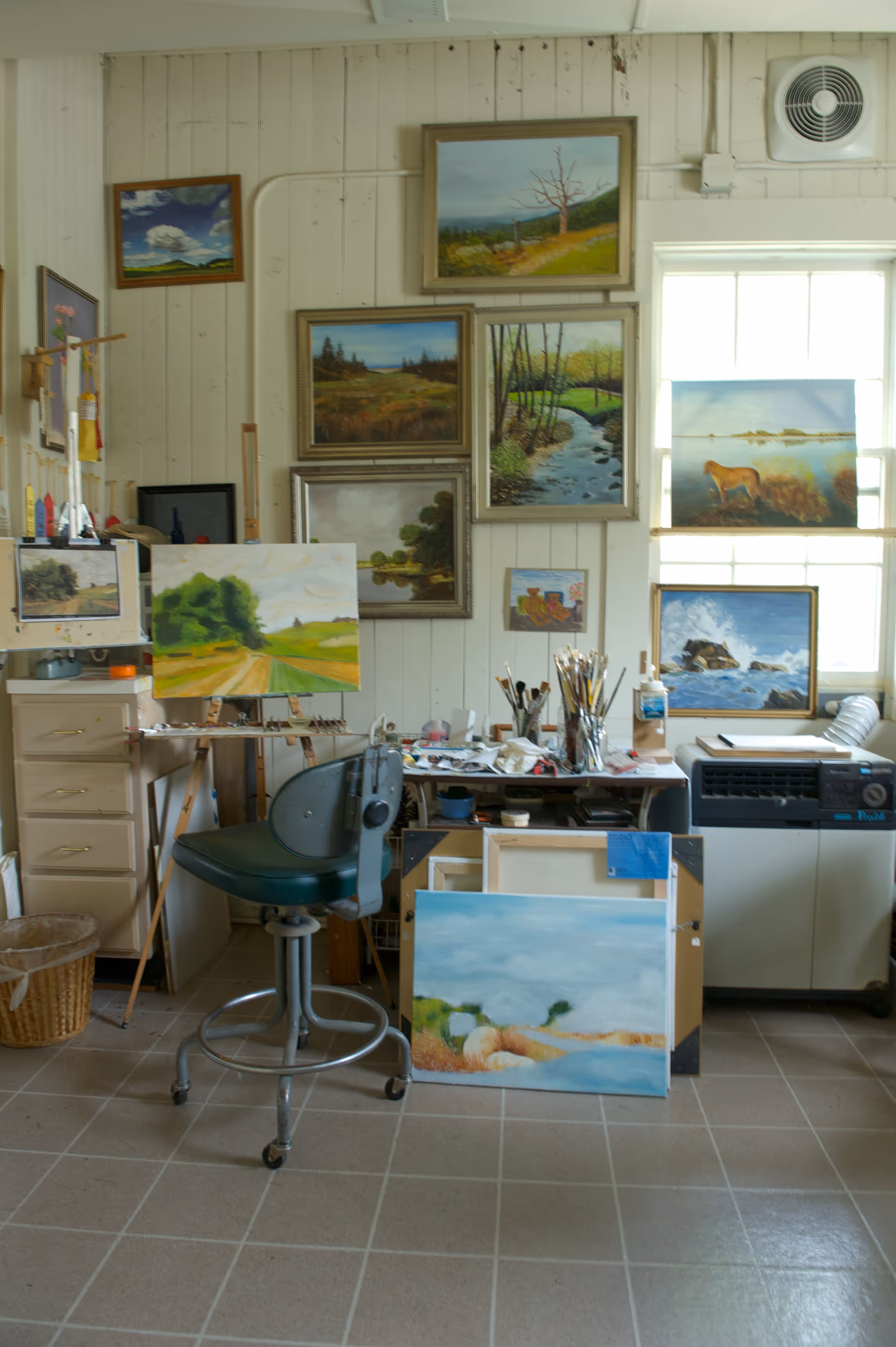 An art studio with multiple landscape paintings displayed on the walls and on easels. There is a green swivel chair in front of a cluttered work table holding paintbrushes and art supplies. The room has tiled flooring, a window letting in natural light, and a ventilation fan on the wall.