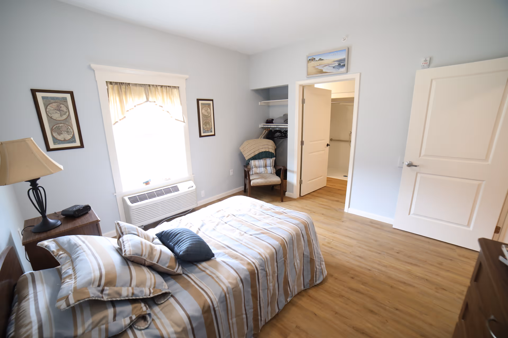 A bright bedroom with a single bed covered in striped bedding and multiple pillows. There is a wooden nightstand with a lamp and a clock radio next to the bed. A window with a light curtain lets in natural light. Two framed pictures hang on the wall beside the window. An open door reveals a bathroom with a shower, and another door is partially open. A chair with folded blankets is placed near an open closet with hangers and shelves. The floor is wooden, and the walls are painted light blue.