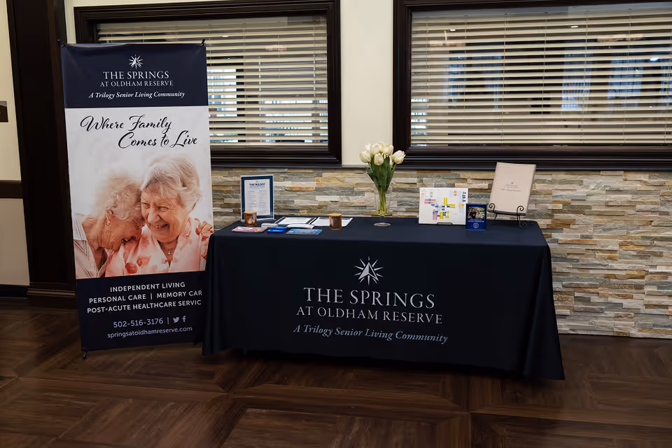 A reception-area display table with brochures, a vase of flowers, and a standing banner for The Springs at Oldham Reserve.