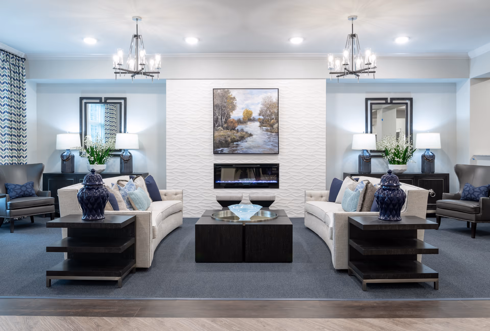 Bright, elegant communal living room with two curved sofas facing a wall-mounted fireplace and artwork, flanked by side tables, lamps, and armchairs.