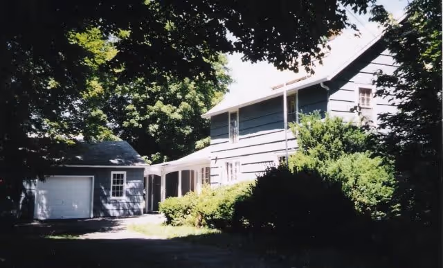 Exterior view of a two-story house with gray siding surrounded by trees and bushes. There is a detached garage to the left connected by a covered walkway. The scene is shaded by large leafy trees.