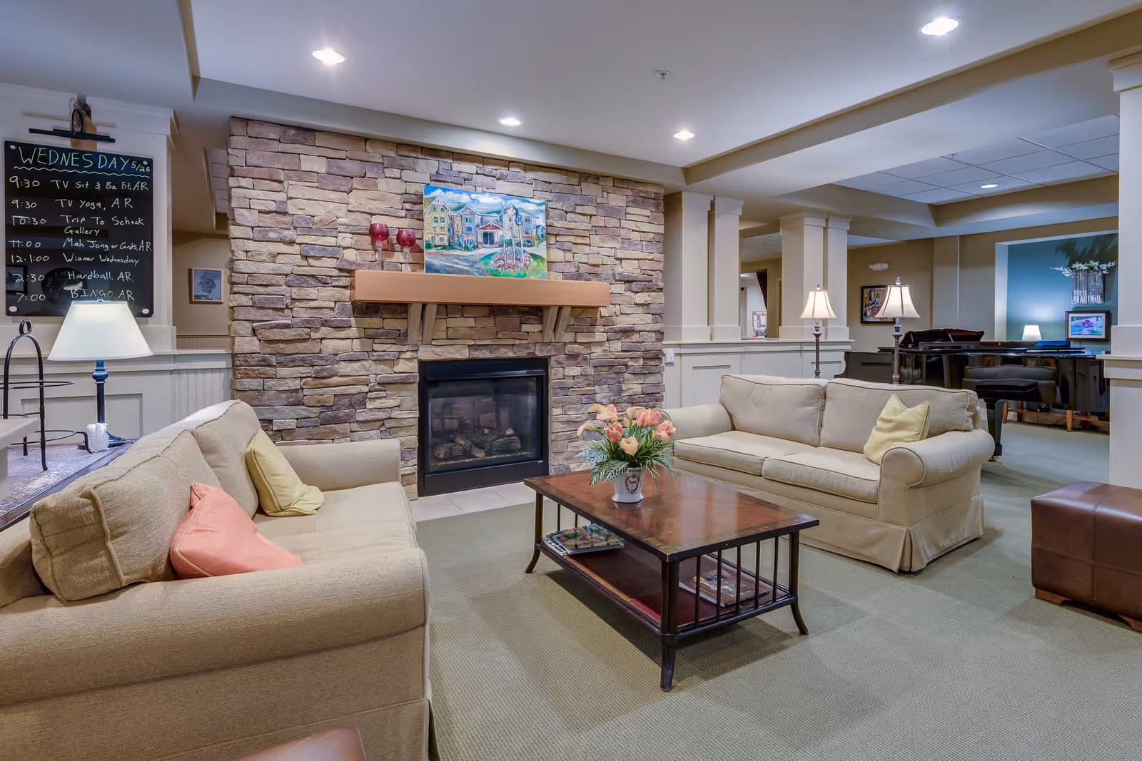 A cozy living room area in a senior living facility with two beige sofas facing each other, a wooden coffee table with a flower vase, and a stone fireplace with a painting above it. There is a blackboard on the left side listing daily activities, and a piano is visible in the background on the right side.