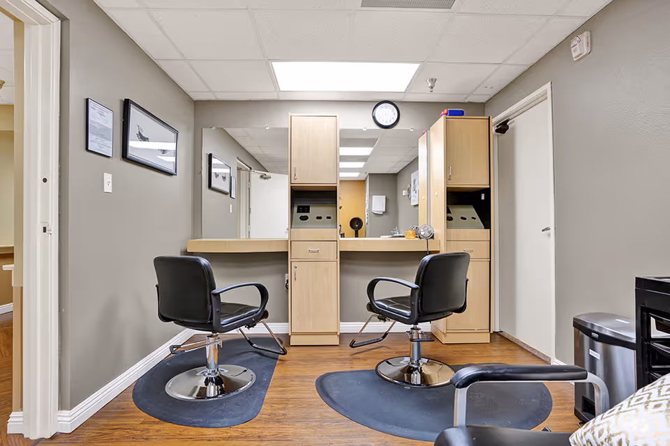 Interior view of a small salon area with two black salon chairs in front of a large mirror mounted on the wall. The mirror is flanked by light wood cabinets with drawers and shelves. The room has gray walls, a wooden floor, and a white ceiling with fluorescent lighting. There is a clock above the mirror and framed pictures on the left wall.