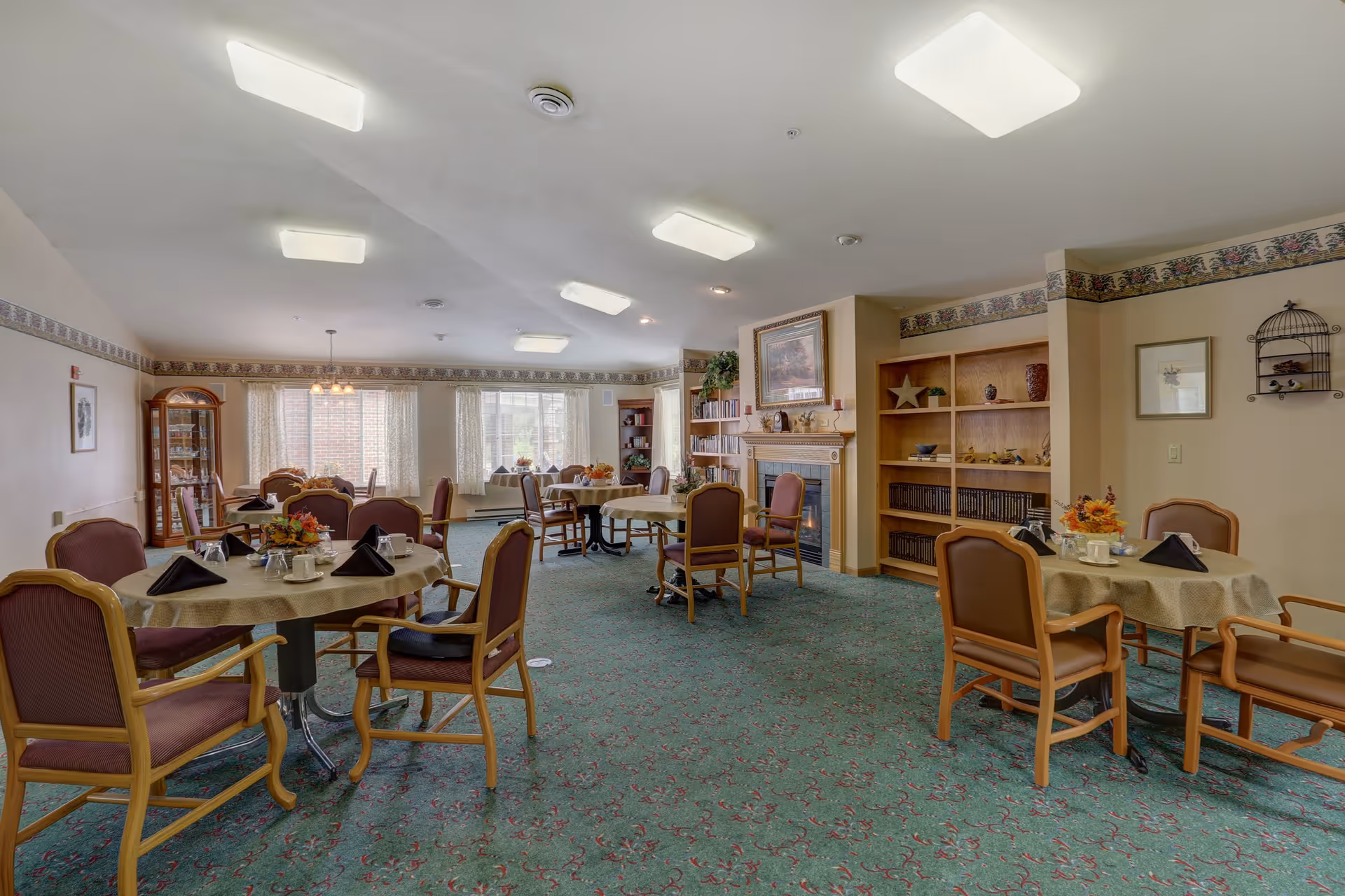 A spacious dining room with several round tables covered with beige tablecloths, each set with black napkins, cups, and floral centerpieces. The room has green patterned carpet, wooden chairs with padded seats, large windows with sheer curtains allowing natural light, a fireplace with a framed picture above it, and built-in wooden shelves with decorative items.