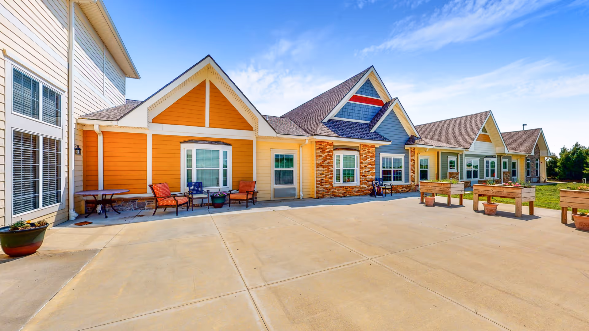Front exterior of a colorful single-story senior living building with a paved courtyard, outdoor seating, and planter boxes under a blue sky.