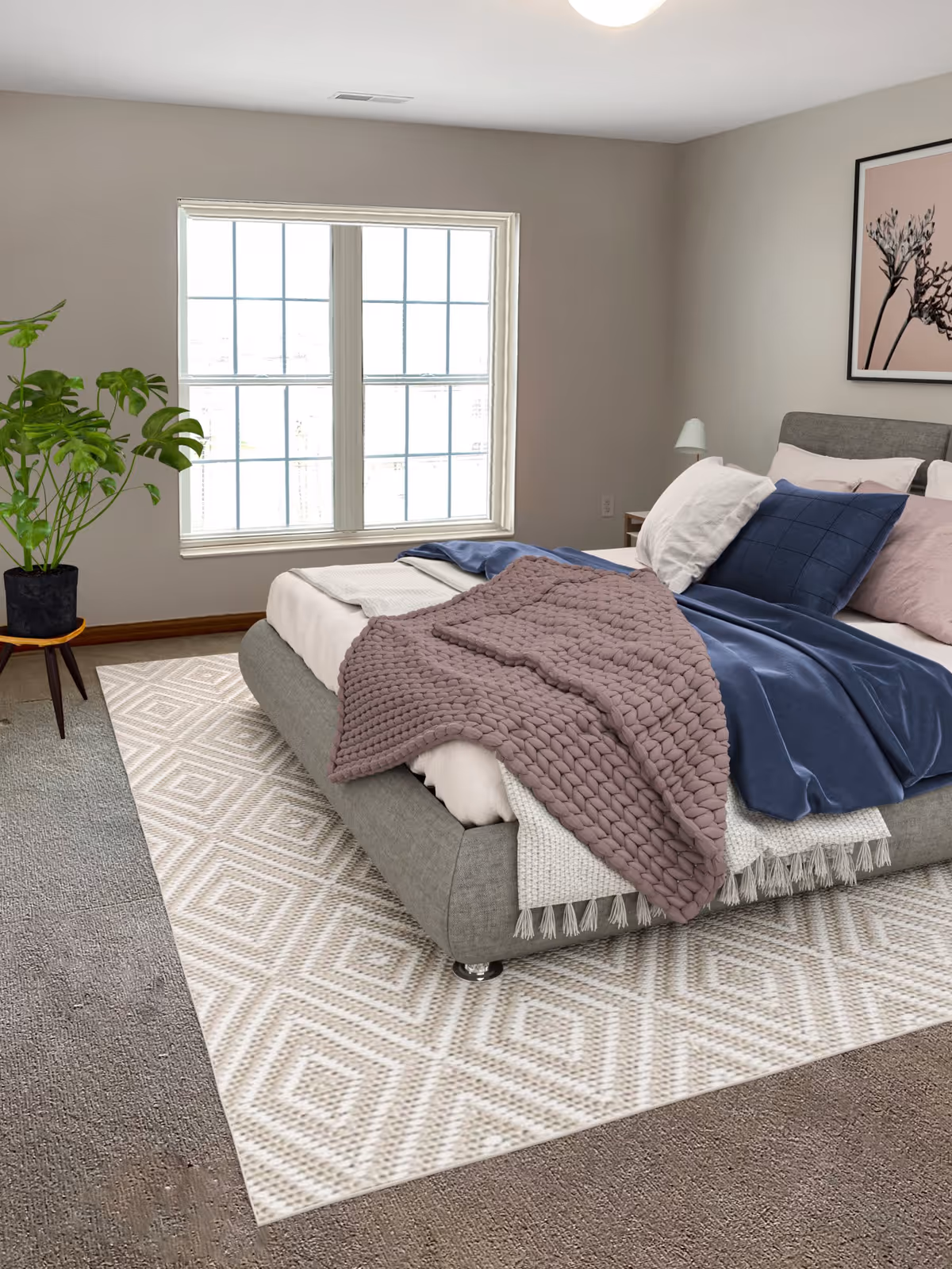 Bright bedroom featuring a gray upholstered bed with layered blankets and pillows, a large window, a patterned rug, a potted plant, and wall art.
