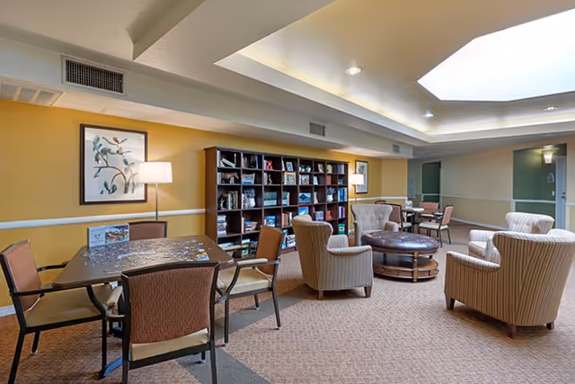 Bright communal lounge with bookshelves, tables, and upholstered chairs under recessed ceiling lighting.