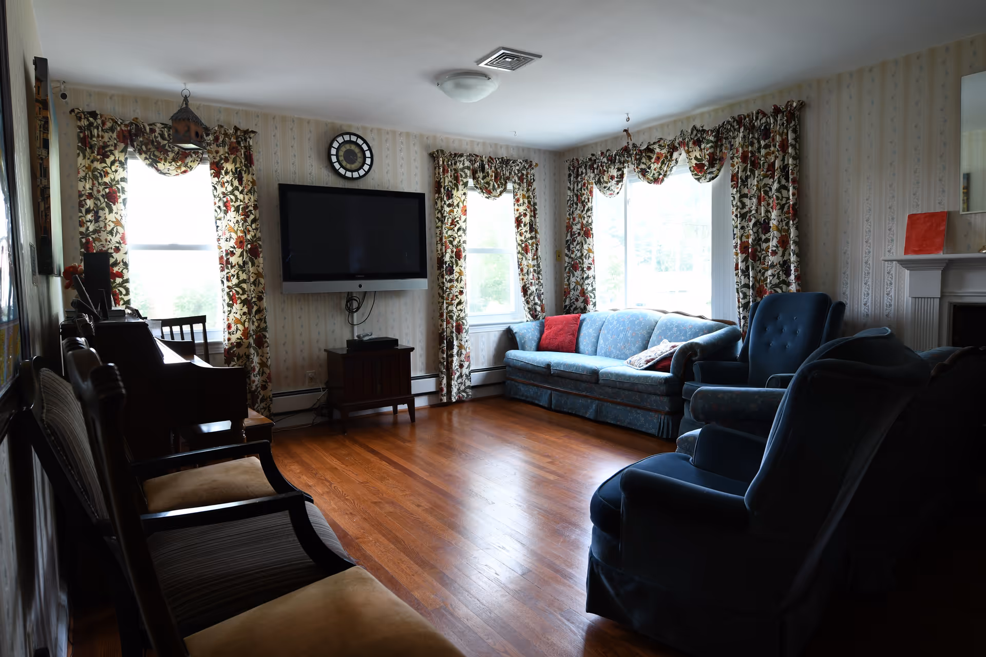 A cozy living room with wooden flooring, floral curtains on three windows, a wall-mounted flat-screen TV, a clock above the TV, a blue sofa with red and patterned cushions, two blue armchairs, a wooden side table, and a fireplace with a red book on the mantel.
