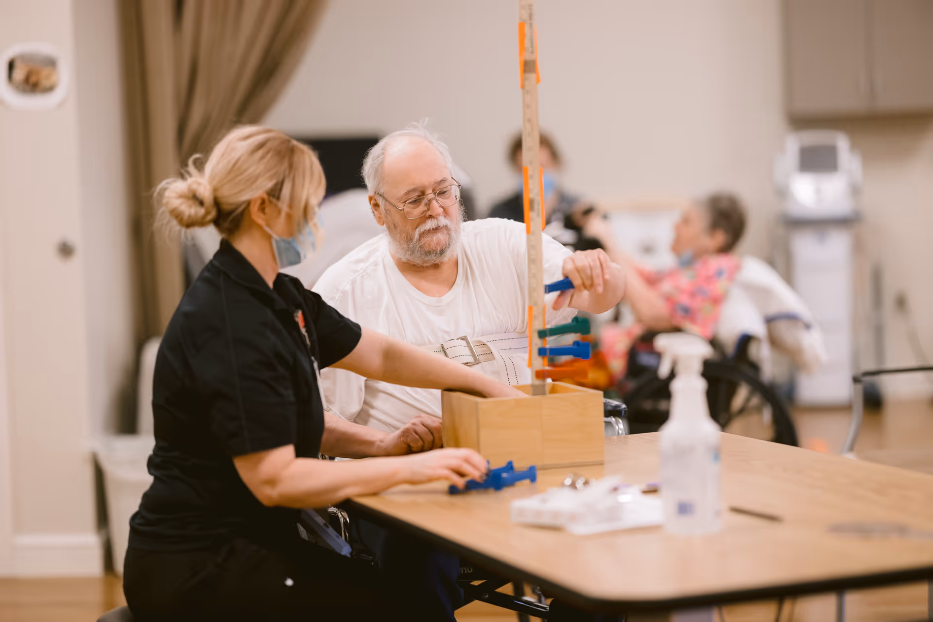 A healthcare worker wearing a mask assists an elderly man with a physical therapy activity involving a wooden pegboard at a table in a care facility. Another elderly person in a wheelchair is visible in the background.