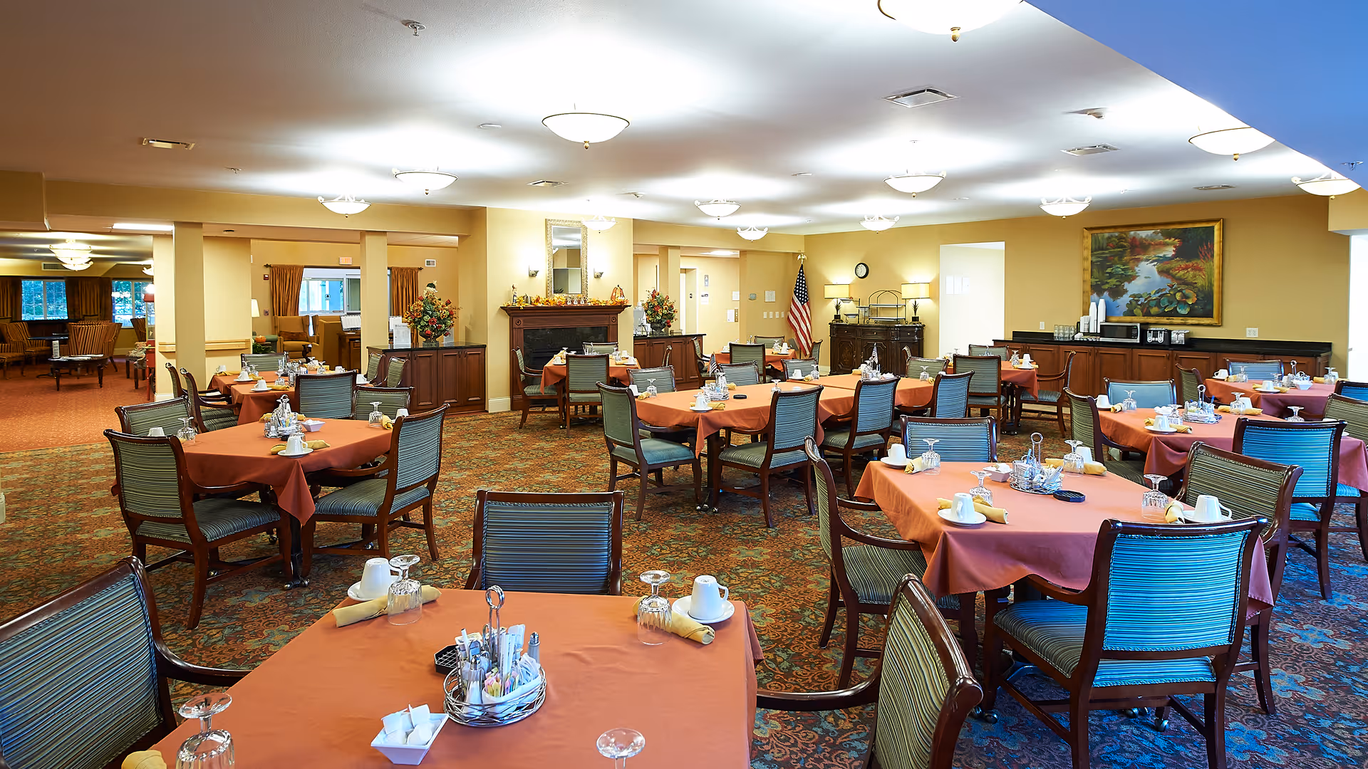 Spacious dining room with multiple round tables covered in rust-colored tablecloths, place settings and upholstered chairs under warm ceiling lights.