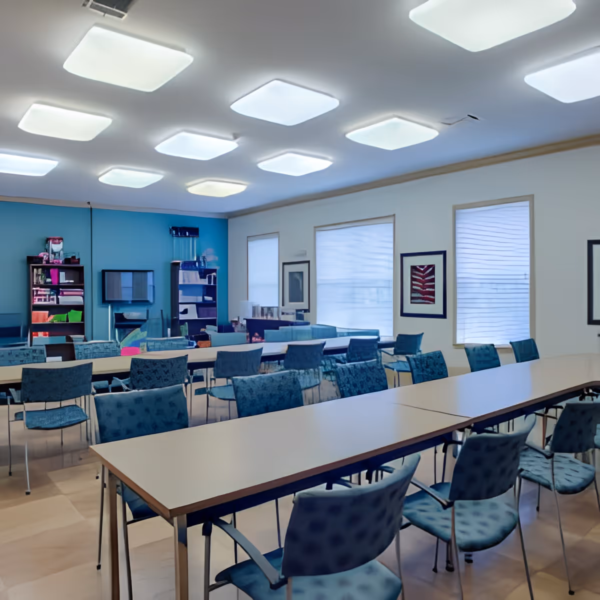 A bright and spacious meeting or activity room with long tables arranged in rows and multiple blue patterned chairs. The room has large windows with blinds, framed artwork on the walls, and modern square ceiling lights. Shelves with various items are visible against a blue accent wall.