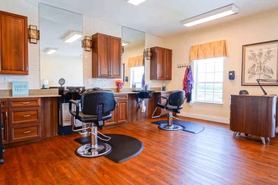Interior view of a hair salon area with two black salon chairs in front of large mirrors mounted on a wall with wooden cabinets. The floor is wooden, and there is a window with a yellow valance letting in natural light. A small wooden desk with a lamp and framed botanical artwork is visible on the right side.