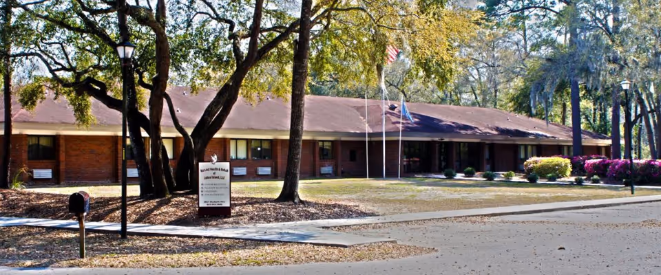 Single-story brick senior living facility with flagpoles, trees, and a manicured lawn.