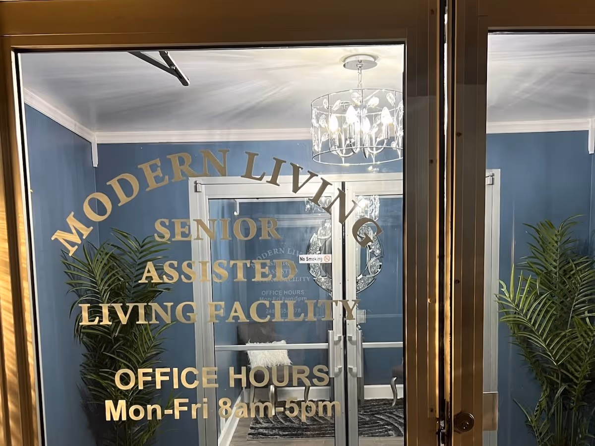 Glass front doors with gold lettering reading 'Modern Living Senior Assisted Living Facility' revealing a blue lobby with plants and a chandelier and posted office hours.