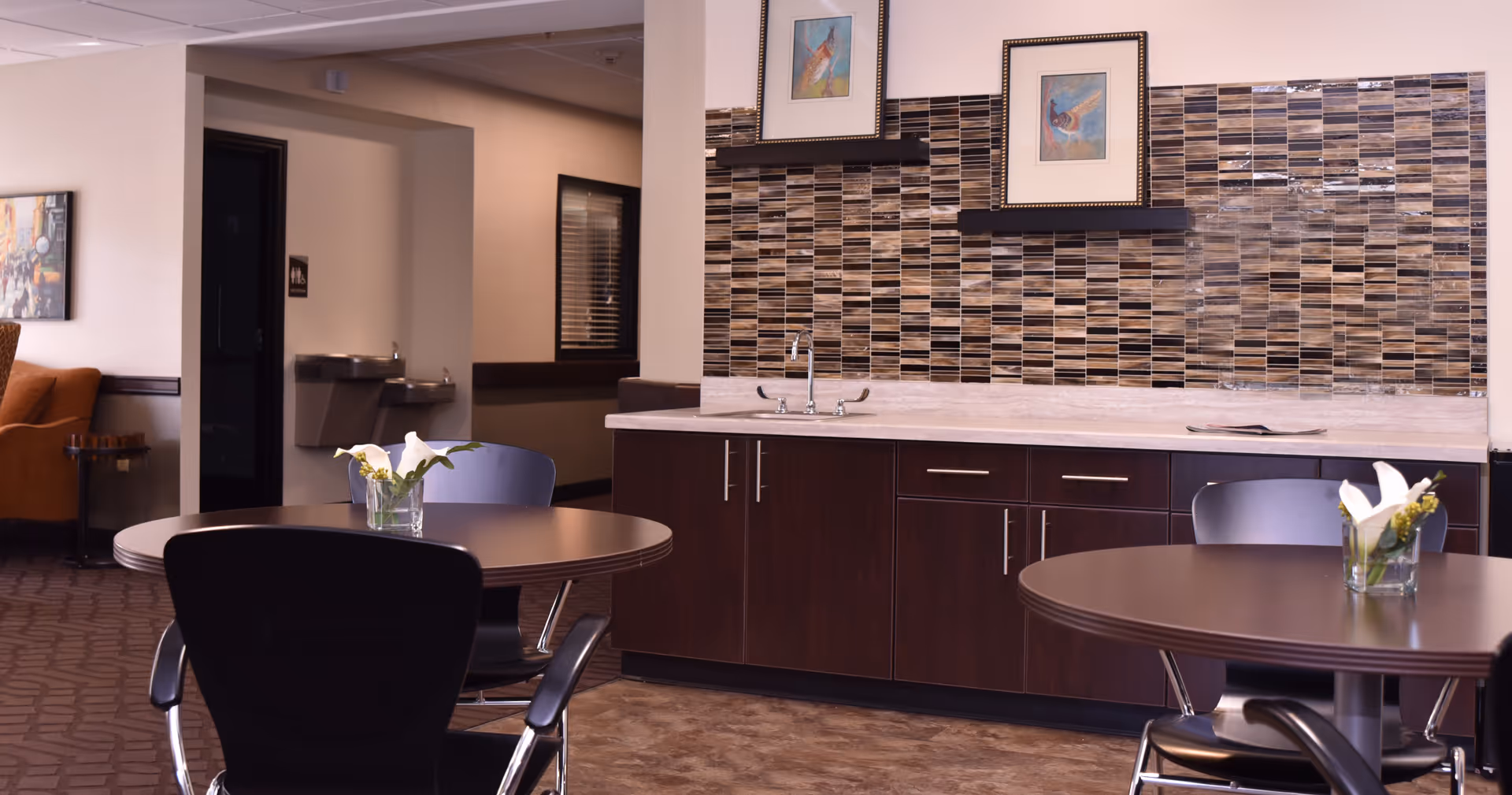 Interior view of a common area in a senior living facility with round tables and chairs, a countertop with a sink, decorative tile backsplash, framed artwork on the wall, and a water fountain in the background.