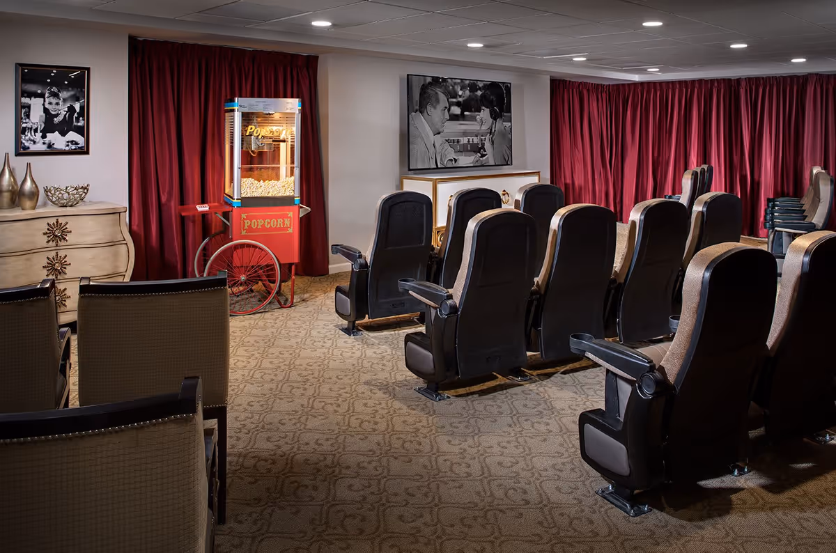 A small theater room with rows of cushioned seats facing a large screen displaying a black and white movie scene. There is a vintage-style popcorn machine on the left side near red curtains, and decorative items including framed black and white photos and vases on a wooden dresser.