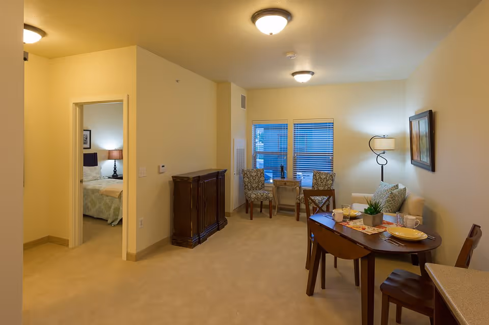 Interior view of a senior living facility apartment showing a small dining area with a wooden table set for two, a cozy sitting area with a couch and two chairs near windows with blinds, and a glimpse into a bedroom with a bed and a bedside lamp.